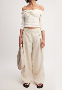 Schouderloze, crèmekleurige top met gedraaide voorkant, gecombineerd met brede, beige linnen broek. Het model houdt een lichtbeige tas vast en draagt zwarte plateau-sandalen.