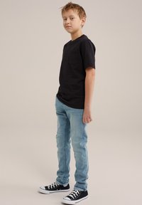 Čierne bavlnené tričko s vreckom, svetlo modré slim-fit džínsy a čierne tenisky s bielymi podrážkami a šnúrkami, stojaci postoj.