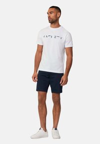 Camiseta de algodón blanca con "HACKETT" impreso en letras oscuras, combinada con pantalones cortos azul marino y zapatillas blancas, sobre un fondo neutro.