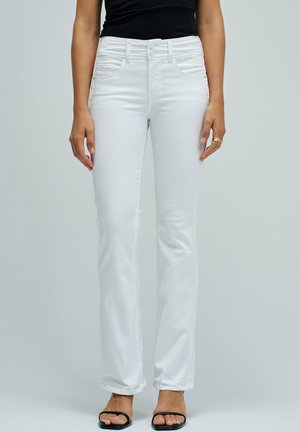 Mujer vistiendo vaqueros blancos de corte bootcut y top negro sin mangas, de pie con los brazos relajados a los lados frente a un fondo liso.