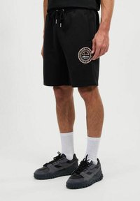 Pantalones cortos negros con cinturilla elástica y cordón. Presenta un parche de logo circular en colores contrastantes. Se lleva con zapatillas negras y calcetines blancos.