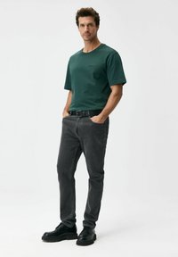 Krótki rękaw ciemnozielonej koszulki z logo, zestawiony z szarymi dżinsami i czarnymi butami na sznurowadła. Prosty fason, casualowy krój i jednolite kolory.