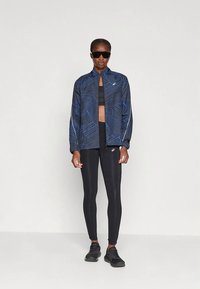 Donna che indossa leggings sportivi neri, reggiseno sportivo nero, giacca zip blu a fantasia, scarpe da ginnastica nere e occhiali da sole scuri, in piedi.