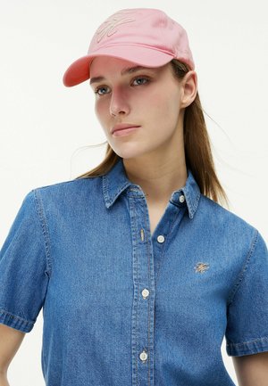 Donna che indossa un berretto da baseball rosa e una camicia di jeans blu a manica corta con le iniziali ricamate sul petto, guarda lontano dalla macchina fotografica.
