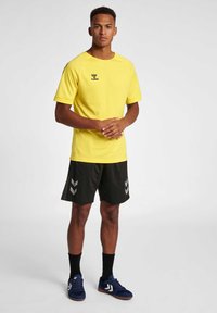 Gul sportstrøje med tekstureret overflade, sorte shorts med sølvfarvede chevronmønstre og blå sneakers med hvide detaljer.