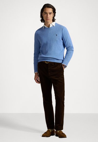 Pull en tricot bleu clair avec des poignets côtelés, associé à un pantalon en velours côtelé marron. Le modèle porte une chemise blanche à col en dessous.