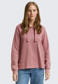 Sudadera con capucha de color rosa claro, con tejido texturizado, ajuste relajado y cordones. Combinada con vaqueros azules, presentada sobre un fondo neutro.