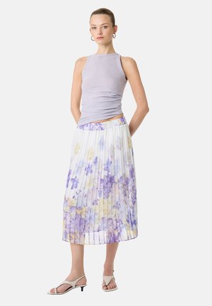 Kvinde iført lavendelfarvet top uden ærmer, hvid plisseret midi-nederdel med lilla og gult blomsterprint samt hvide åbne kitten heels.