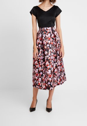 Femme vêtue d'un haut noir à manches courtes et d'une jupe midi taille haute à motif floral, portant des escarpins noirs.