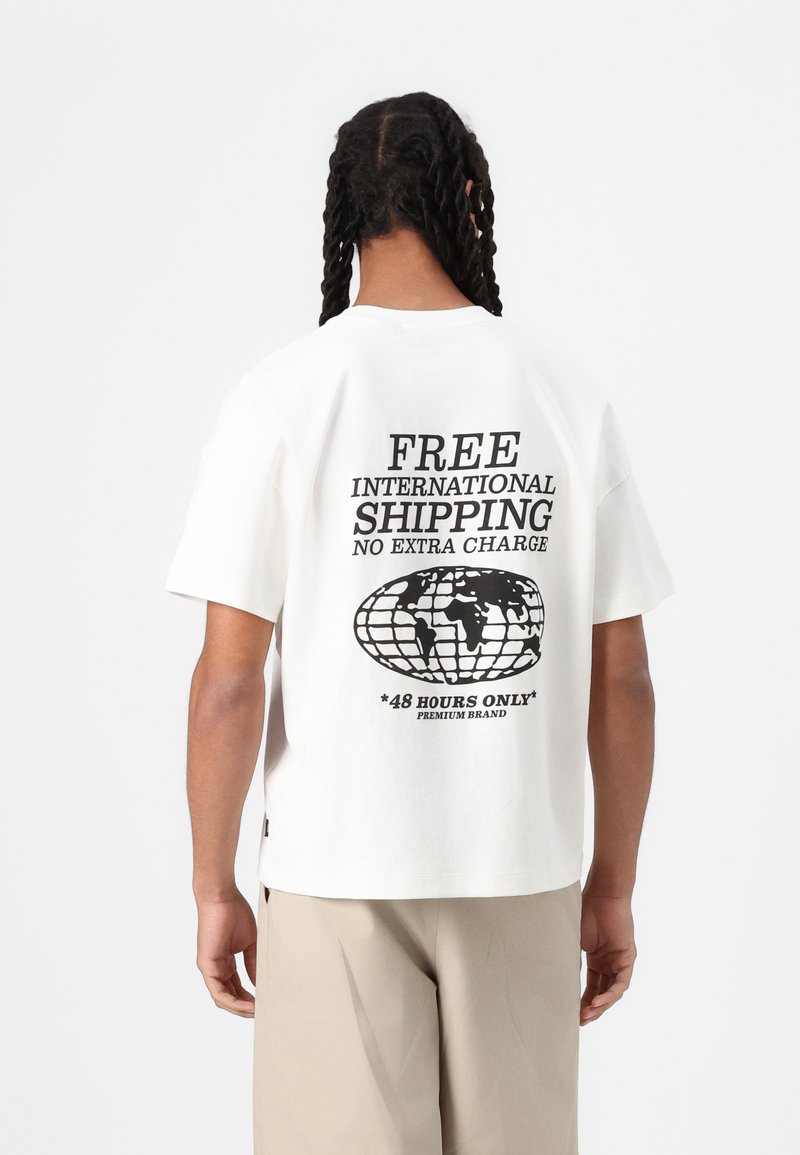 T-shirt in cotone bianco con una grande grafica nera sul retro che include testo riguardante la spedizione e un design del globo. Vestibilità rilassata e maniche corte.
