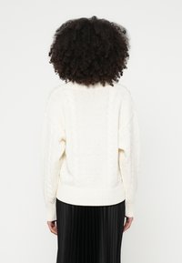 Maglione lavorato a maglia color avorio con motivo a trecce, polsini e orlo a coste, con dettagli del logo "HUGO" tessuto. Abbinato a una gonna nera plissettata.