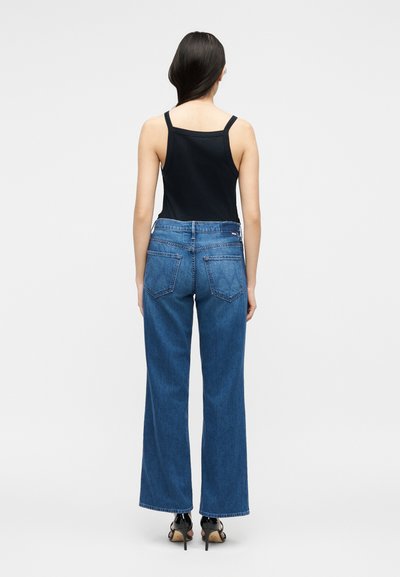 Γυναίκα που φαίνεται από πίσω, φοράει μαύρο αμάνικο τοπ, μπλε τζιν με φαρδιά πόδια και μαύρα πέδιλα με ψηλό τακούνι σε απλό φόντο.