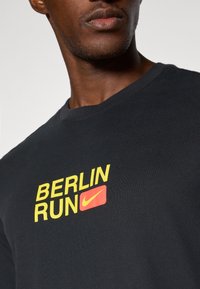 Black cotton t-shirt featuring "BERLIN RUN" in yellow text and a red Nike logo. Round neckline and short sleeves.