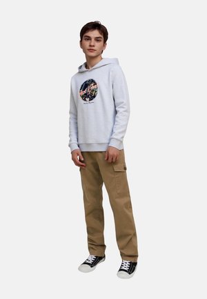 Heller grauer Hoodie mit grafischem Druck, beige Cargo-Hosen, schwarze Sneakers mit weißen Sohlen; das Modell steht mit entspannten Händen an den Seiten.