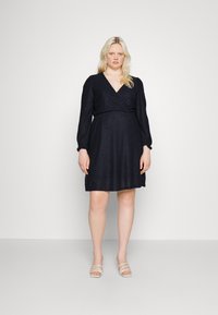 Vestido azul-marinheiro em formato envolvente, com mangas bouffant longas, tecido texturizado e um laço na cintura. Apresenta um decote em V e uma bainha evasê, combinado com sandálias de cor clara.