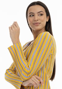 Femeie cu păr lung și închis la culoare, îmbrăcată cu un pulover în dungi galbene, mov și maronii, privindu-se din lateral cu brațele încrucișate.
