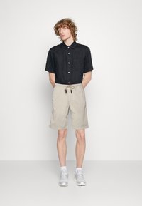 Short-sleeve black button-up shirt paired with beige drawstring shorts. Model wears gray sneakers, standing against a plain background.