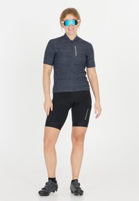 Cykeltröja i mörkgrått med texturerad tyg, korta ärmar och dragkedja framtill. Svarta cykelshorts med vadderad insats. Blå spegelglasögon.