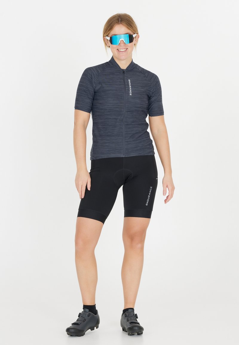 Cykeltröja i mörkgrått med texturerad tyg, korta ärmar och dragkedja framtill. Svarta cykelshorts med vadderad insats. Blå spegelglasögon.