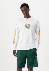 Long-sleeve white shirt featuring flame graphics and a logo on the chest, paired with green patterned shorts and a small logo accent.