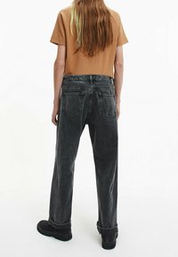 Osoba z długimi brązowymi włosami, ubrana w brązową koszulę, czarne jeansy z podwiniętymi nogawkami i czarne buty, stojąca tyłem.