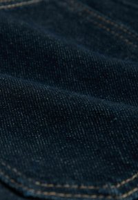 Donker denim stof met een gladde textuur. Zichtbare stiksels in lichtbruin en subtiele kleurvariaties door het materiaal.