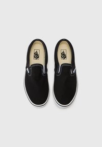 Fekete vászon slip-on cipők fehér varrással és gumitalppal. A belső szegmens bézs talpbetéttel rendelkezik, amelyen a logó látható. Tiszta, minimalista dizájn.