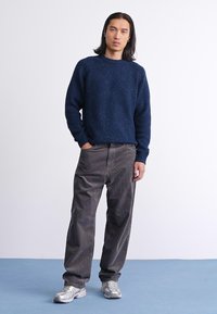 Granatowy sweter z dzianiny o strukturalnej fakturze, w zestawieniu z szarymi spodniami z korduroy. Model nosi srebrne buty sportowe, pozując na jednolitym tle.
