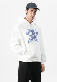 Sudadera blanca con texto azul que dice "TOMMY ATTEMPT JEANS NEW YORK." Presenta un bolsillo delantero, cordones y puños acanalados. Combinada con pantalones negros.