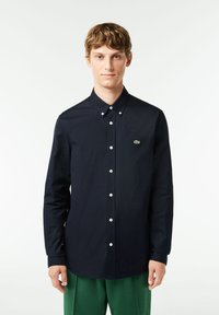 Camicia blu navy con bottoni, maniche lunghe, bottoni bianchi e un piccolo logo verde sul lato sinistro del petto, realizzata in un tessuto liscio.