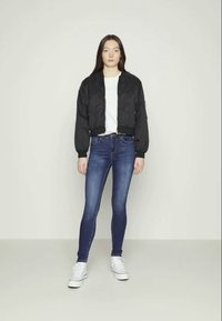 Jaqueta bomber preta com textura acolchoada, t-shirt branca, jeans skinny azul escuro e ténis brancos. Modelo está posicionado contra um fundo simples.