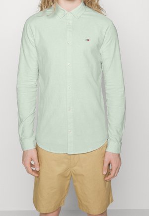 Persona con camisa verde claro de botones y pantalones cortos beige, de pie contra un fondo liso, con las manos relajadas a los lados.