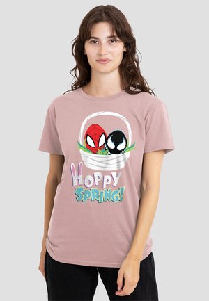 Frau trägt ein mauvefarbenes T-Shirt mit einem Osternest, in dem Spider-Man- und Venom-Eier abgebildet sind, sowie dem Schriftzug "Hoppy Spring!" vor einem schlichten Hintergrund.