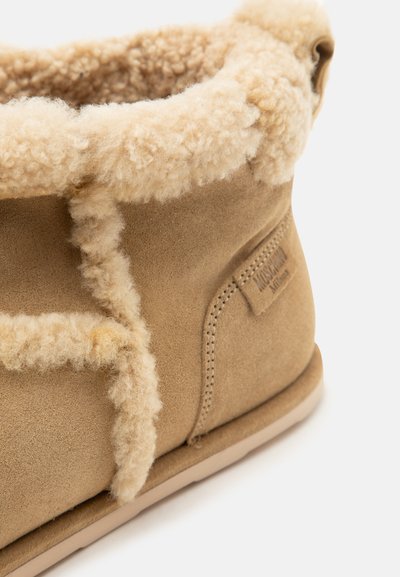 Gros plan d'une botte d'hiver en daim beige avec une doublure et une finition en peau de mouton crème, montrant la couture latérale et une partie de la semelle.