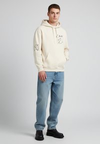 Felpa crema con grafiche di uccelli neri sulle maniche, tasca a marsupio frontale, abbinata a jeans azzurri chiari e stivaletti neri.