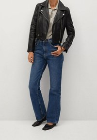 Persoon in een zwarte leren cropped jack, beige polo, blauwe high-waist rechte jeans en zwarte puntige ballerina's, staande tegen een witte muur.