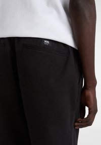 Black pants made from smooth fabric, featuring side pockets and a visible Vans logo patch on the waistband. Hand resting on the fabric.