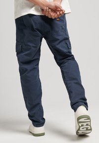 Pantalones cargo de color azul marino con bolsillos laterales, corte relajado y detalle de logo bordado. Combinados con zapatillas blancas con acentos verdes en la suela.