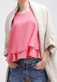 Haut sans manches rose à plusieurs couches en tissu doux, associé à un jean en denim bleu clair. Un cardigan beige texturé complète la tenue.