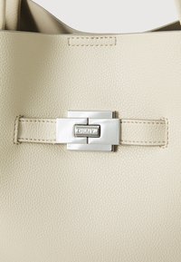 Beige skinnveske med en strukturert overflate, med en sølvfarget metallklips med logo og en sydd stroppdetalj.
