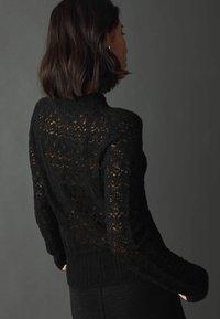 Maglione nero a maglia con un design a rete lavorata, dotato di maniche lunghe e orlo a coste, evidenziando il contrasto di texture e l'artigianato.
