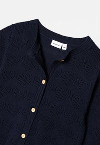 Marineblauer Cardigan mit Herzmuster, ausgestattet mit rundem Ausschnitt, Knopfverschluss und hellen Holzknöpfen. Hergestellt aus strukturiertem Strickmaterial.