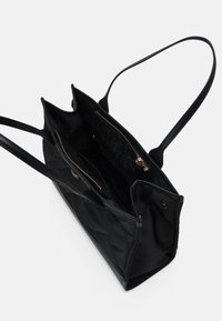 Sac tote noir en matériau lisse et matelassé. Comprend deux compartiments zippés et de longues poignées fines. Intérieur avec impression de logo.