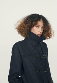 Il cappotto in lana navy presenta un collare alto, chiusure a toggle e un tessuto testurizzato con grandi tasche rettangolari sul davanti.