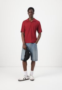 Red ribbed shirt with short sleeves, denim shorts, white and gray sneakers, holding a black bag. Plain background.