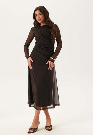 Kvinna poserar i en midjelång svart klänning med genomskinliga långa ärmar, rynkad midja, pärlarmband och leopardmönstrade öppna klackskor mot vit bakgrund.