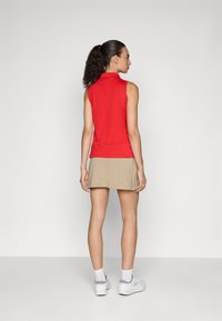 Rød ermeløs polo med høy krage, parret med en beige skjørt. Modellen har hestehale og bruker hvite joggesko med grå detaljer.