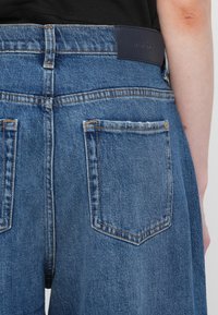Blå denimjeans har høy midje, baklommer, kontrasterende oransjefargede sømmer og et mørkeblått lærmerke på livet.