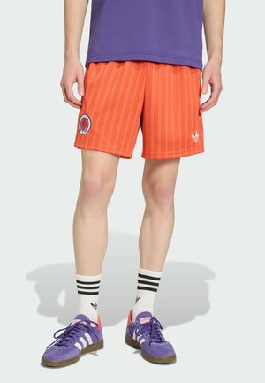 Modell trägt orange gestreifte Shorts mit Logos, weiße Socken mit schwarzen Streifen und violette Sneakers mit weißen Streifen, steht vor einem schlichten Hintergrund.