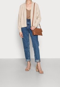 Beige cardigan over a brown blouse, high-waisted blue denim jeans, and tan block-heeled sandals. Brown crossbody bag with braided detail.